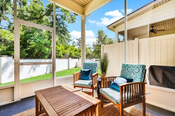 A covered patio area with a wooden table and two chairs with patterned cushions, a potted plant, and a view of a green lawn and trees.