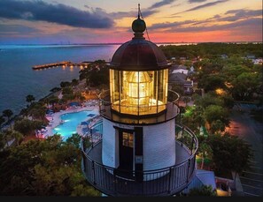 A lighthouse with a glass enclosure, surrounded by a pool and lounge area, overlooking a coastal landscape.