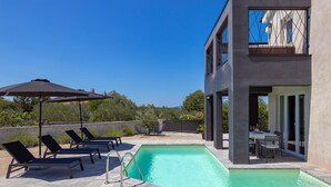 A modern outdoor pool area with lounge chairs and a dining table, surrounded by a landscaped garden and a clear blue sky. - Villa Nika by Interhome (Krk/Dobrinj)