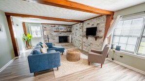 A spacious living room with a brick fireplace, a television mounted on the wall, and a large window with blinds. - Beach Daze Duplex - Unit B! Fenced-in backyard! Pets Welcome! 5 Blocks to beach! (Carolina Beach)