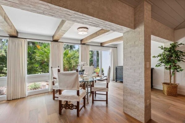 A dining area with a glass table, wooden chairs, and a view of greenery outside.