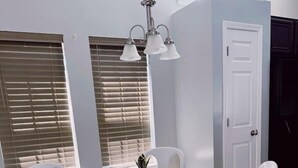 A dining area with a glass table surrounded by white chairs with patterned cushions, a hanging light fixture, and a door in the background. - Coastal Oasis: Games, Pool, BBQ & beach (Myrtle Beach)
