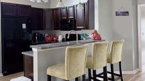 A modern kitchen with dark wood cabinets, a stainless steel refrigerator, a black microwave, and a granite countertop. There are three bar stools with yellow upholstery and pendant lights above the island. - Coastal Oasis: Games, Pool, BBQ & beach (Myrtle Beach)
