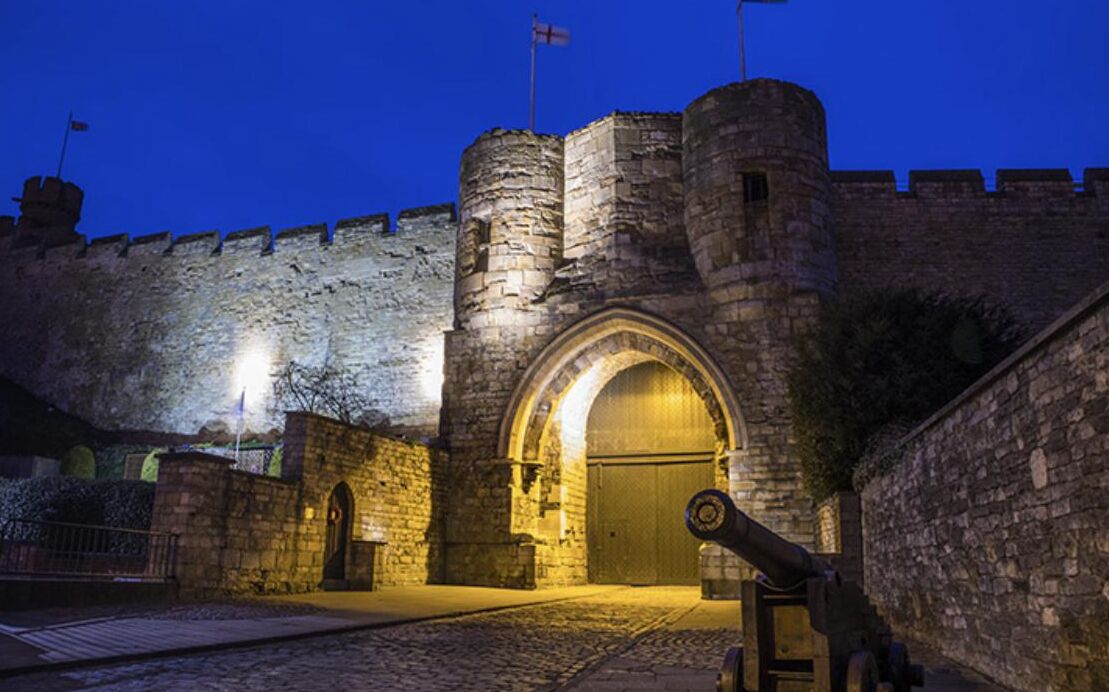 A historic stone castle with a large arched entrance, a flag on top of a tower, and a cannon in front.