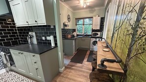 A kitchen with white cabinets, a black countertop, a wooden bar counter, and a window with a view of trees. - Cosy and secluded brook-side lodge (Sleaford)