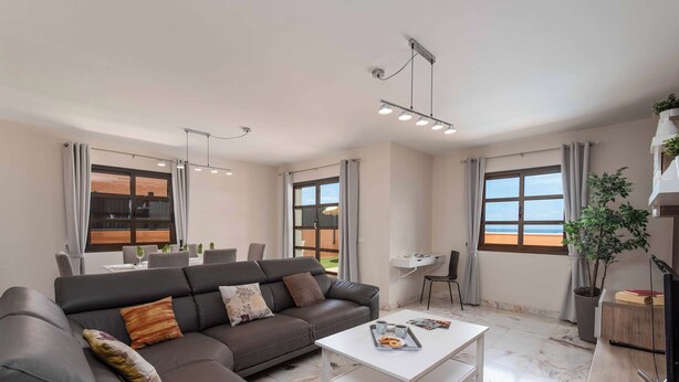 A modern living room with a sectional sofa, a white coffee table, and a dining area with a table and chairs. There are large windows with curtains, a ceiling light fixture, and a potted plant. - Vista Lobos (La Oliva)