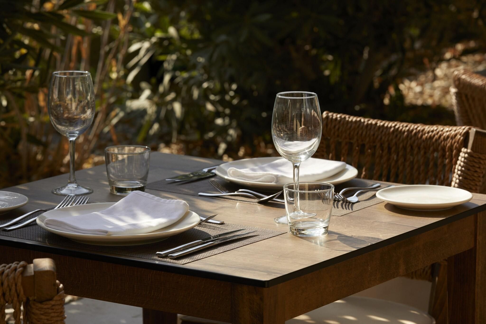 An outdoor dining table set with plates, cutlery, and glasses, surrounded by wicker chairs.