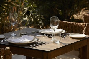 Mesa de comedor al aire libre con platos, cubiertos y vasos, rodeada de sillas de mimbre.