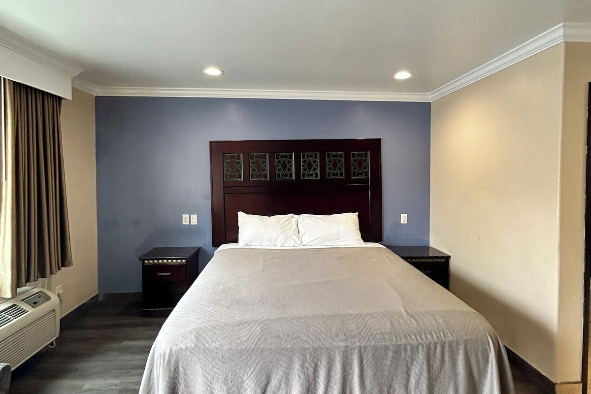 A hotel room with a large bed, a wooden headboard, a nightstand, a wall-mounted heater, and a window with curtains.