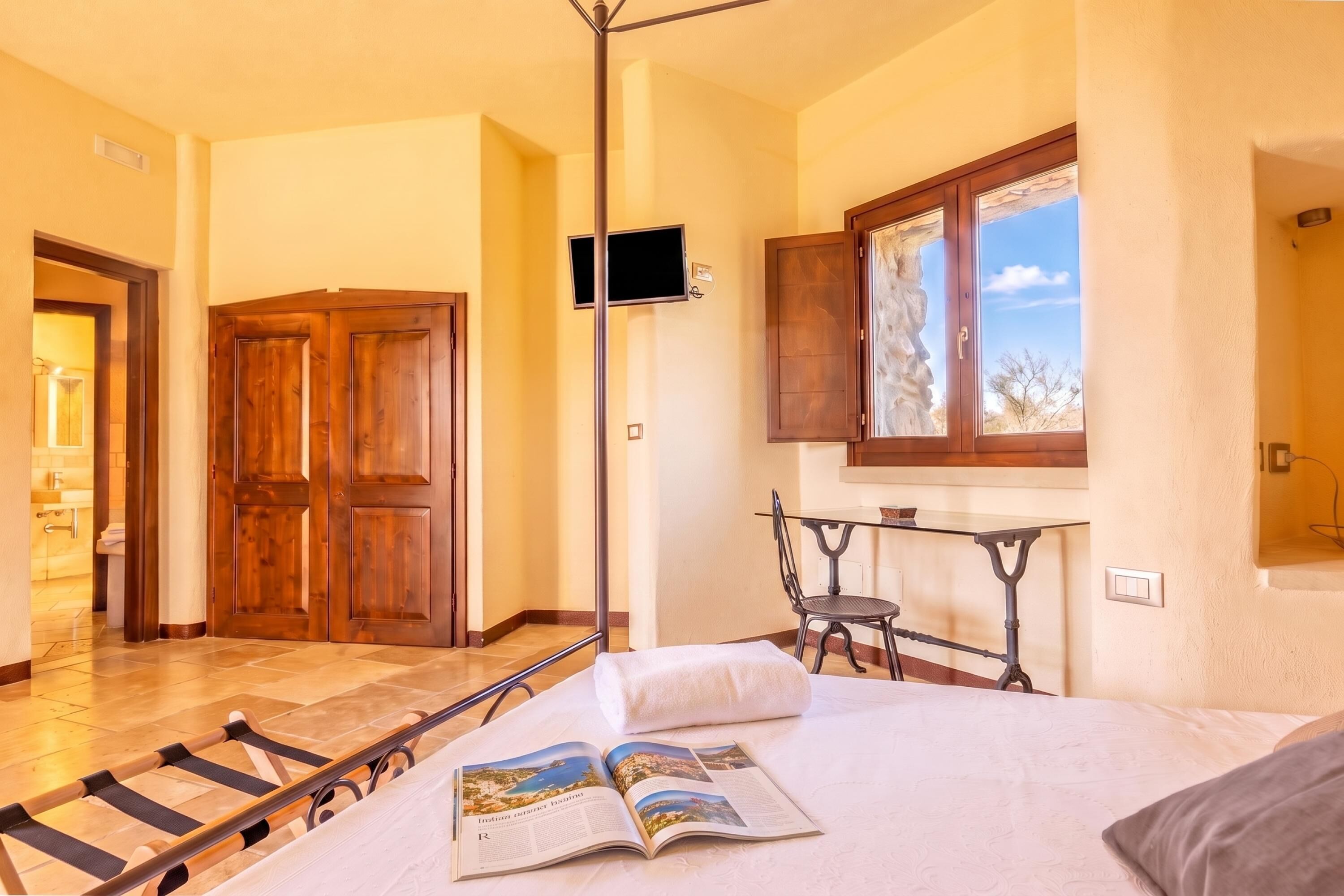A hotel room with a bed, a reading area with a magazine, a window with a view, and a wooden door.