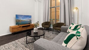 A modern living room with a sofa, armchairs, a coffee table, a TV on a wooden stand, and a large window with curtains. - 1 BR Apartment In the Heart of Darling Harbour (Pyrmont)