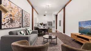 A modern living room with a grey sofa, two armchairs, a coffee table, and a TV on a wooden stand. There are abstract paintings on the wall and a cityscape painting on the TV stand. - 1 BR Apartment In the Heart of Darling Harbour (Pyrmont)