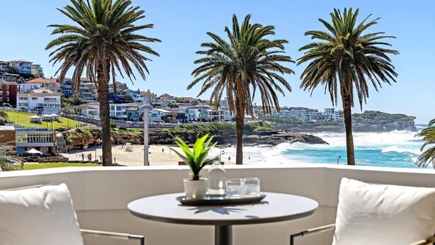 Un balcón con una mesa redonda y dos sillas, con vista a una playa con palmeras y un pueblo costero.