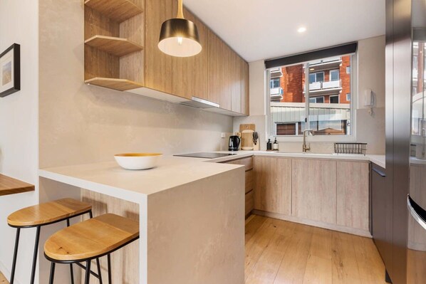 A modern kitchen with wooden cabinets, a white countertop, a built-in island with stools, and a stainless steel refrigerator.