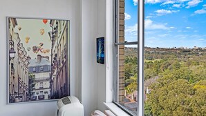 A room with a large window, a painting of a cityscape, and a view of a green landscape. - Centennial Park Light Filled Modern Studio (Centennial Park)
