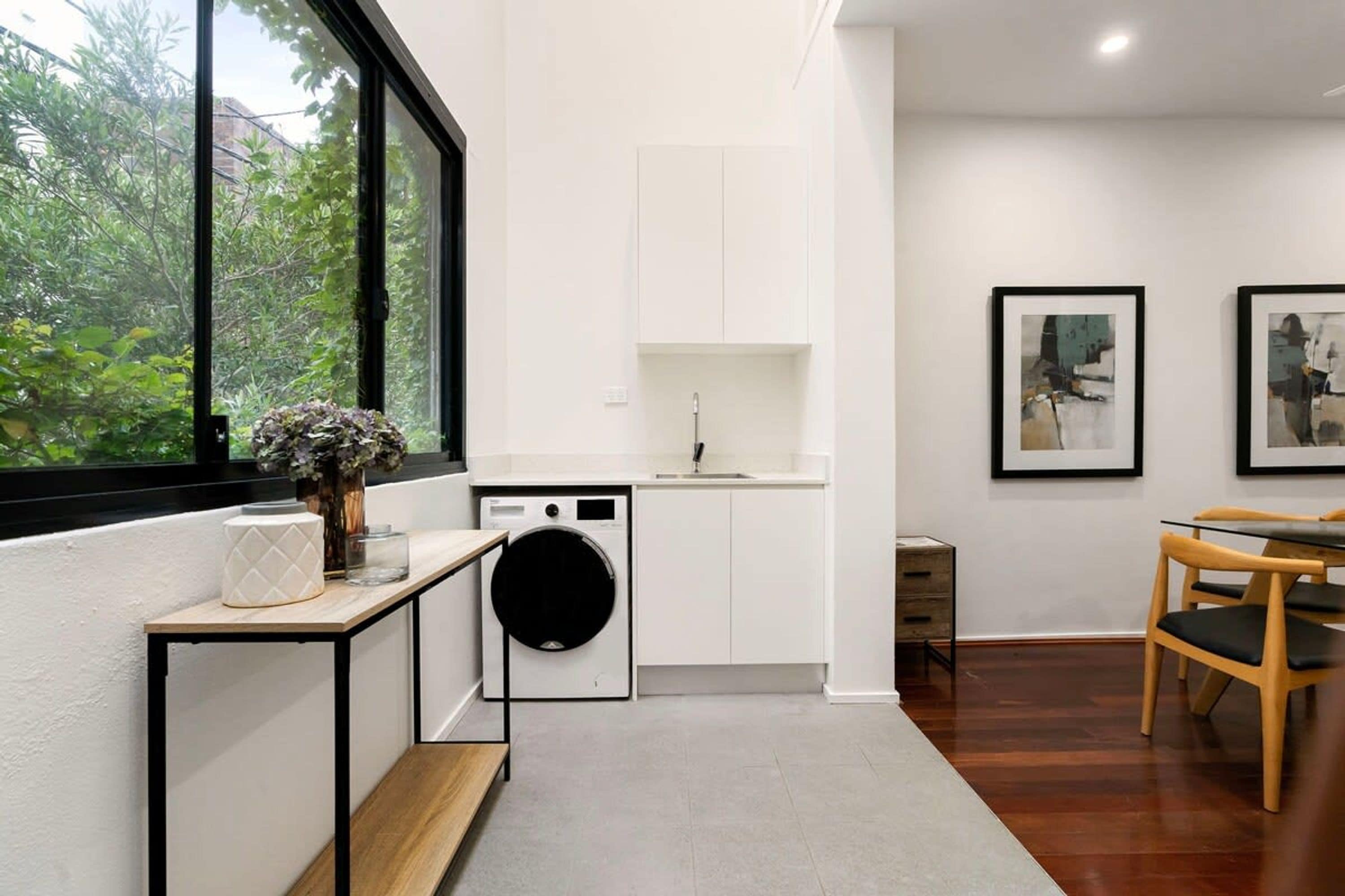 A compact kitchen with a built-in washer, a dining area with a wooden table and chairs, and a large window with a view of greenery.