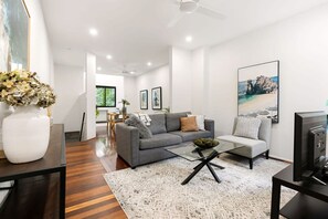Ein modernes Wohnzimmer mit grauem Sofa, einem Couchtisch und einem Fernseher auf einem Schrank. An der Wand hängt ein Gemälde und auf einem Tisch steht eine Blumenvase.