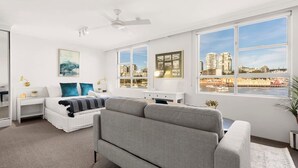 Un salon moderne avec un canapé, un lit agrémenté d’oreillers bleus, un bureau blanc et de grandes fenêtres donnant sur la ville.
