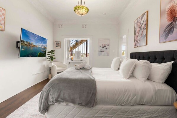 A bedroom with a large bed, a desk, and a chair. There are paintings on the walls and a window with curtains.
