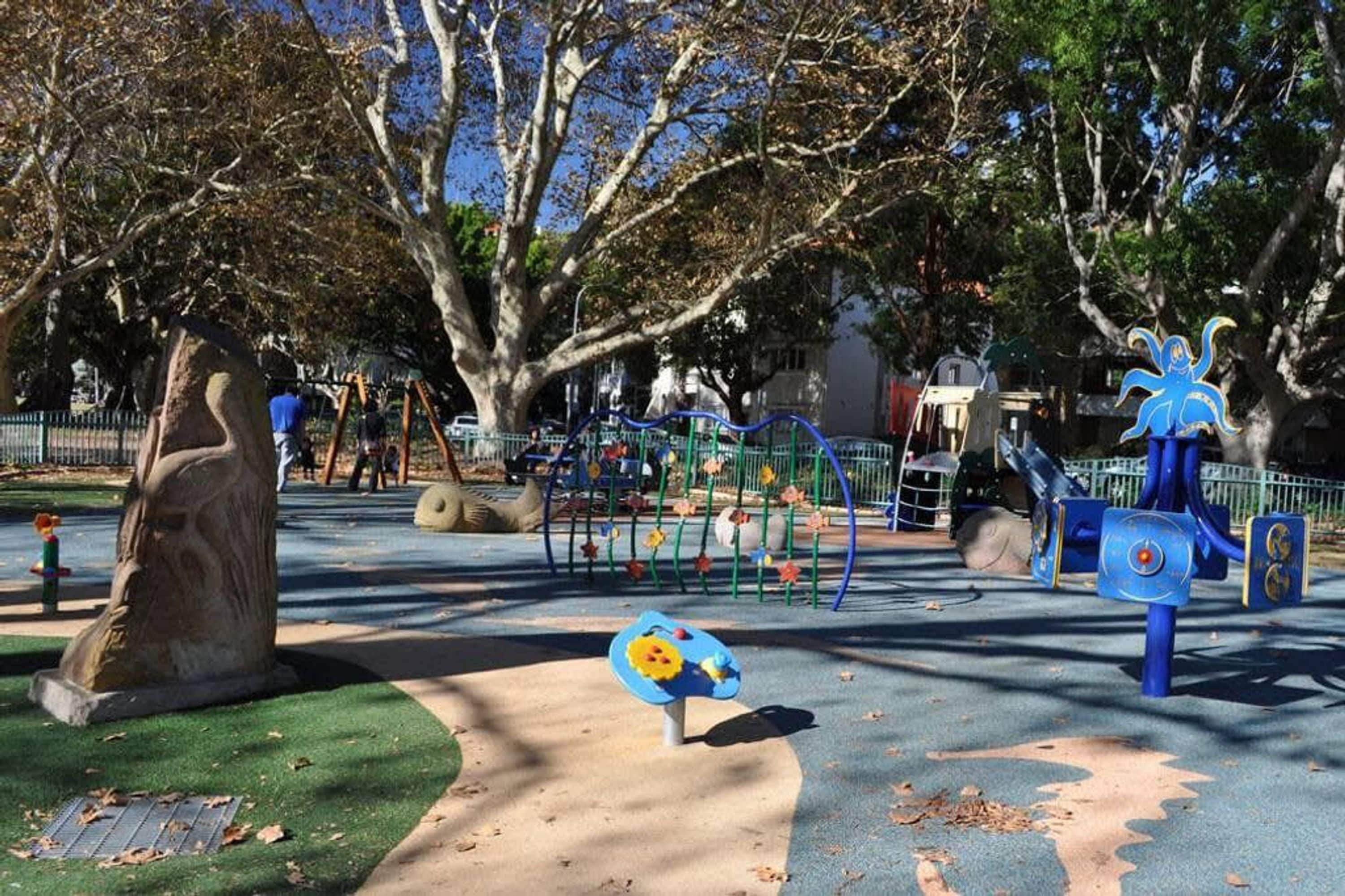 A playground with various play structures, a large tree, and a paved area with a blue and yellow play figure.