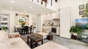 A modern living room with a sofa, coffee table, and dining area. There is a kitchen with white cabinets and a dining table. - Poolside Apartment Opposite Redfern Park (Redfern)