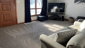 Een woonkamer met een grote flatscreen televisie, een houten deur, donkere gordijnen, een plafondventilator en een schilderij aan de muur.