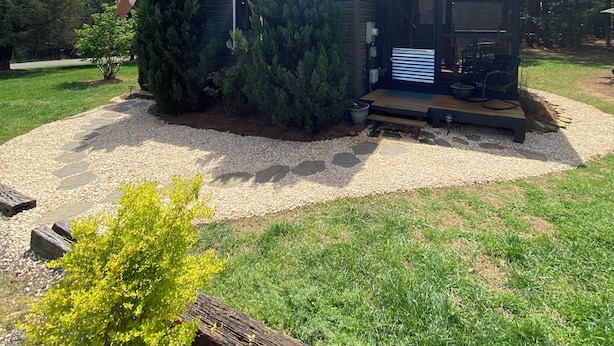 A modern cabin with a deck and outdoor seating, surrounded by a gravel path and greenery.