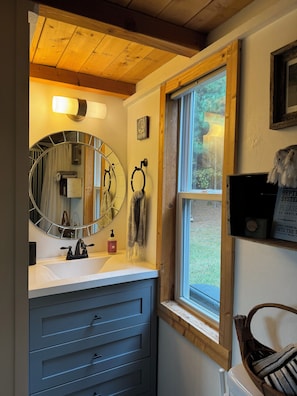 A bathroom with a round mirror, a sink with a soap dispenser, and a window with a view of greenery. - Adventurous tiny house! Located on 1 acre surrounded by evergreen forest (Statesville)