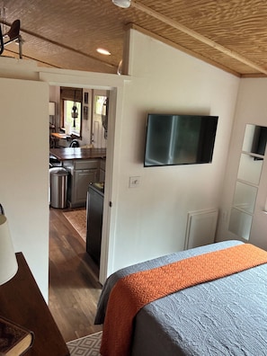 A bedroom with a bed, a television mounted on the wall, and a view into the kitchen area. - Adventurous tiny house! Located on 1 acre surrounded by evergreen forest (Statesville)
