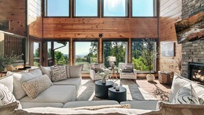 A spacious living room with a large wooden ceiling, a fireplace, and a view of a forested area.