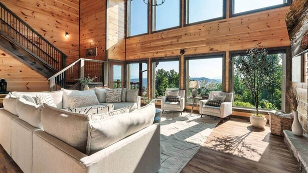 A spacious living room with a high ceiling, wooden beams, a large sofa, two armchairs, and a view of the outdoors.