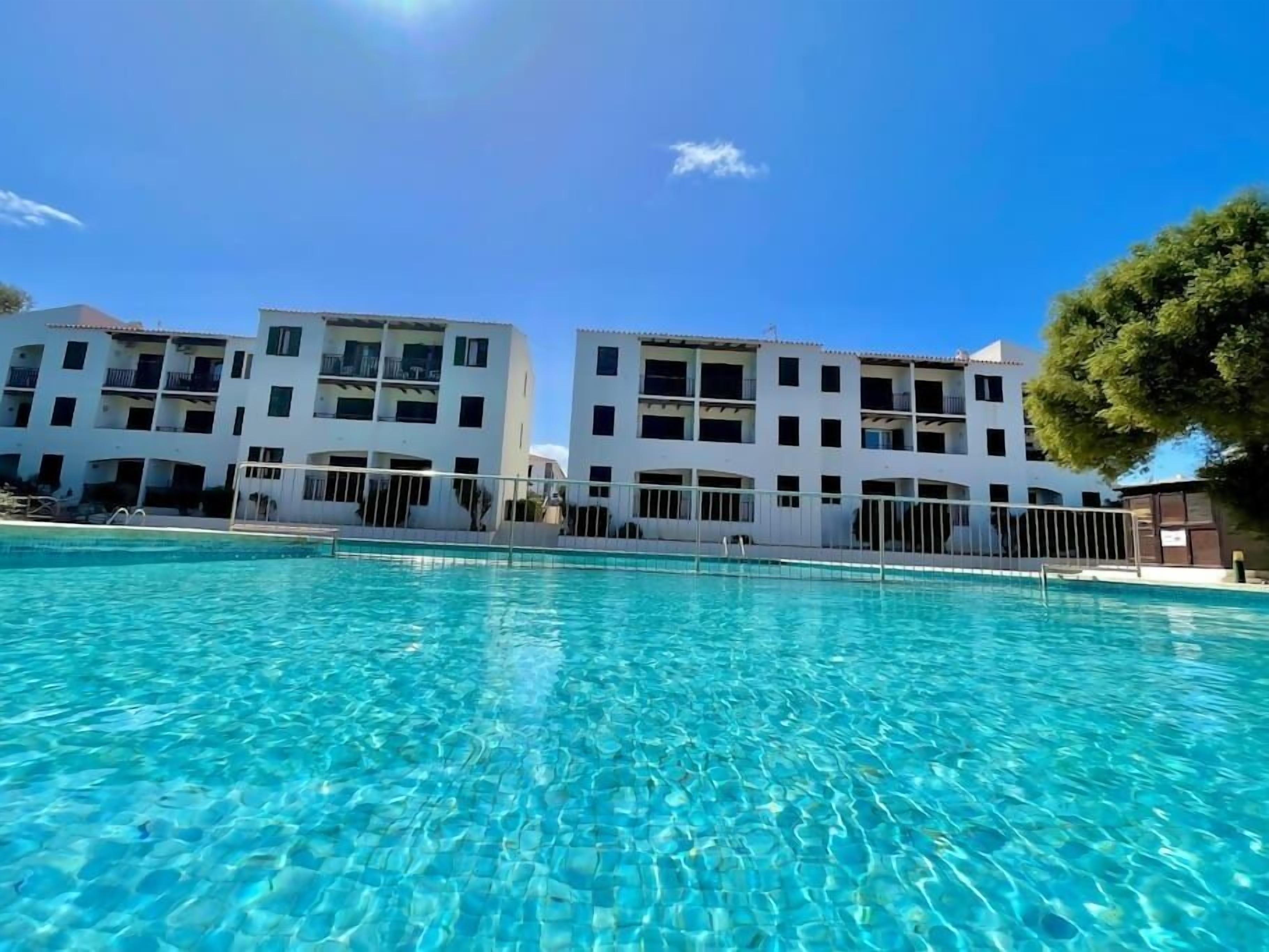 A swimming pool with clear blue water, surrounded by white buildings with balconies and a tree to the right.