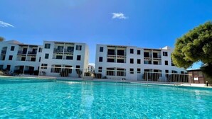 A swimming pool with clear blue water, surrounded by white buildings with balconies and a tree to the right. - Xaloc , Apartamento con Piscina y Junto la Playa (Na Macaret (Urbanitzacio))