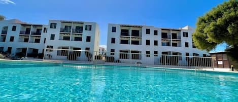 A swimming pool with clear blue water, surrounded by white buildings with balconies and a tree to the right.