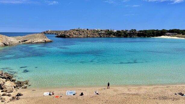 A coastal beach with clear turquoise waters, rocky formations, and a small settlement on a cliff. - Xaloc , Apartamento con Piscina y Junto la Playa (Na Macaret (Urbanitzacio))