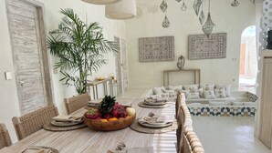 A dining area with a long table set for four, surrounded by wicker chairs. A large bowl of fruit is placed on the table, and there are two wall-mounted light fixtures above. - Casa Blanca (Canggu)