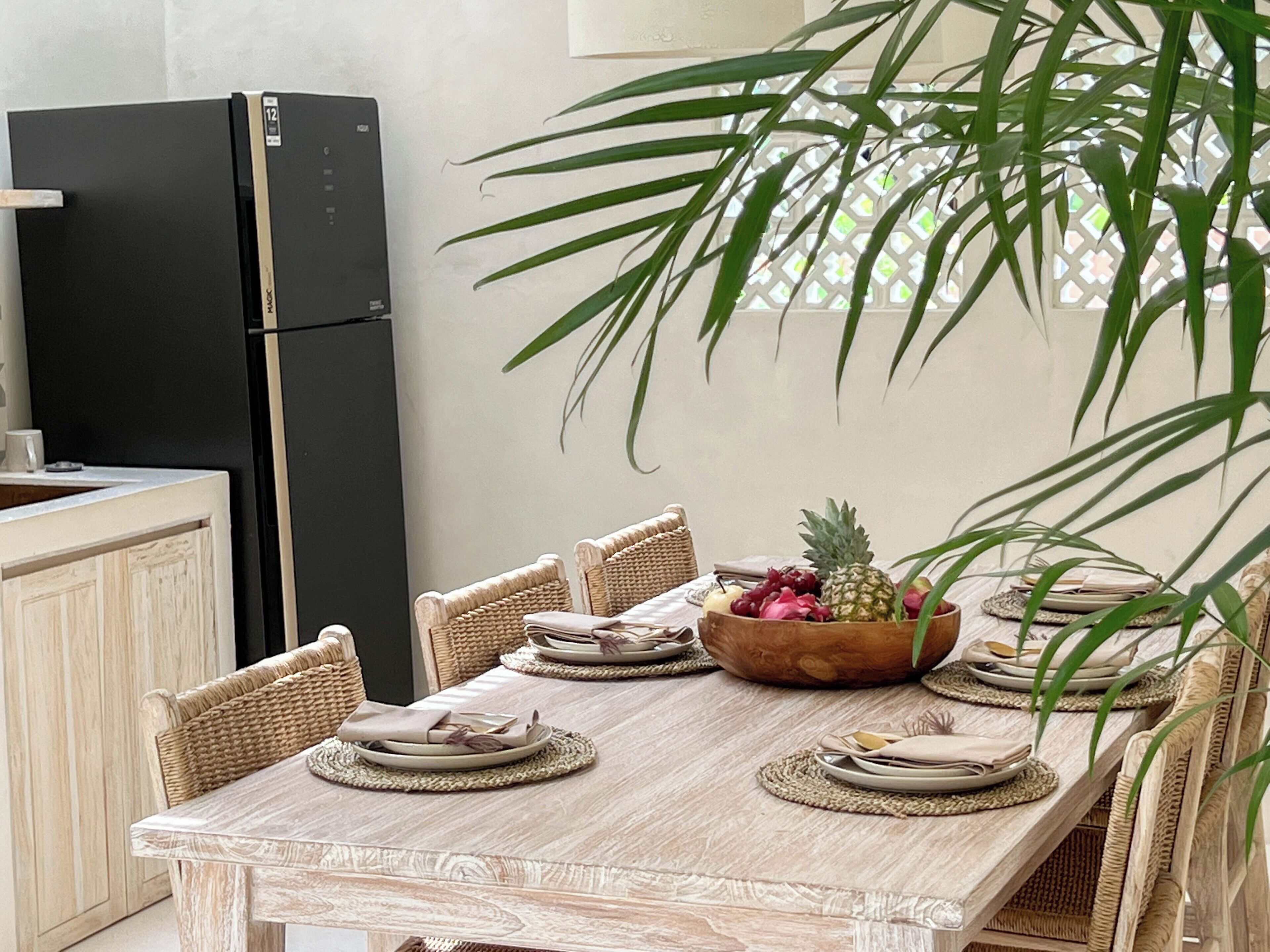 A dining area with a wooden table set for four, surrounded by wicker chairs. A black refrigerator is visible to the left.