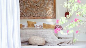 A bedroom with a large decorative panel above the bed, a hanging plant, and a window with curtains. - Casa Blanca (Canggu)