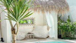 A poolside area with a palm tree, striped cushions, a small table with a teapot, and a thatched roof. - Casa Blanca (Canggu)