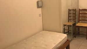 Une petite chambre avec un lit simple, un banc en bois, une chaise et une cloison en verre.
