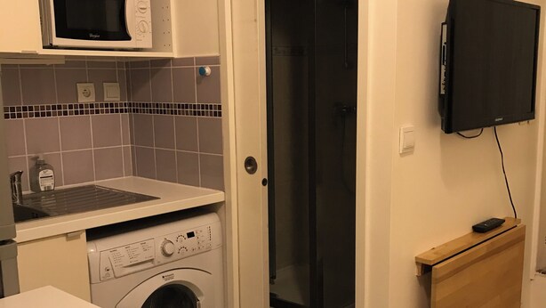 Une cuisine compacte équipée d’un four à micro-ondes, d’un lave-linge et d’un petit plan de travail. On aperçoit une douche derrière une porte ouverte, et un téléviseur à écran plat est fixé au mur.