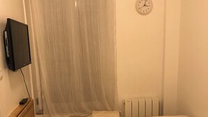 Une petite chambre propre, avec un lit, une table de chevet, un téléviseur, un radiateur et une horloge au mur.