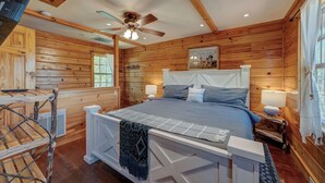 Une chambre à coucher dans une cabane en bois, avec un lit, des lampes de chevet, un ventilateur de plafond et une fenêtre avec des rideaux.