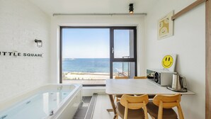 Una habitació moderna amb banyera, taula de dinar amb cadires, finestra gran amb vista al mar i paret amb un rètol que diu 'Little Square'.