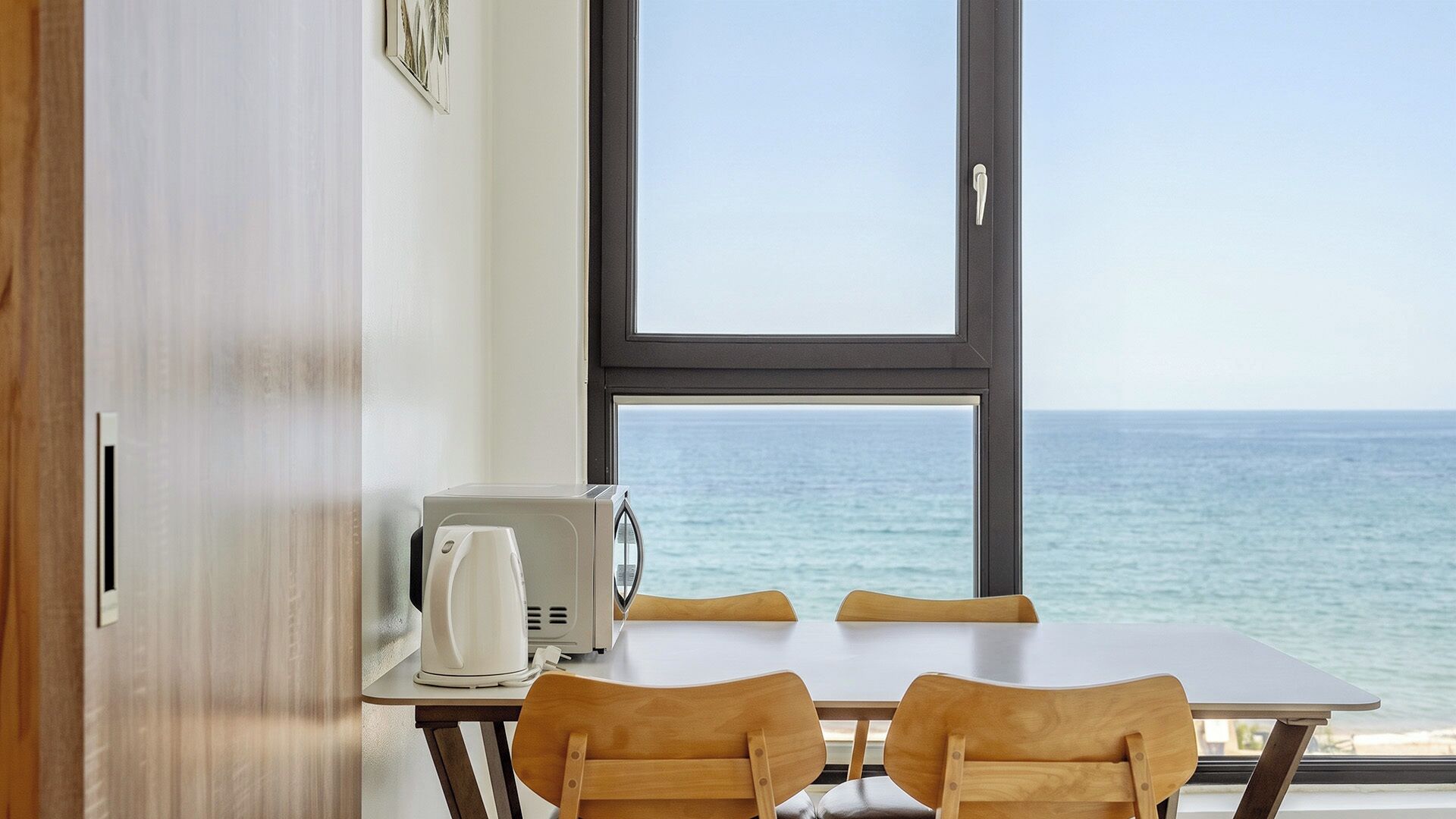 A room with a table and chairs, a coffee maker, and a view of the sea.