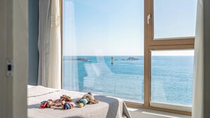 Habitació amb una finestra gran davant d'un mar tranquil, un llit amb edredó blanc i una taula petita amb joguines.
