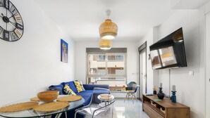 Un salon moderne avec un canapé bleu, une table basse en verre et des chaises transparentes. Une grande horloge et un téléviseur à écran plat sont fixés aux murs.