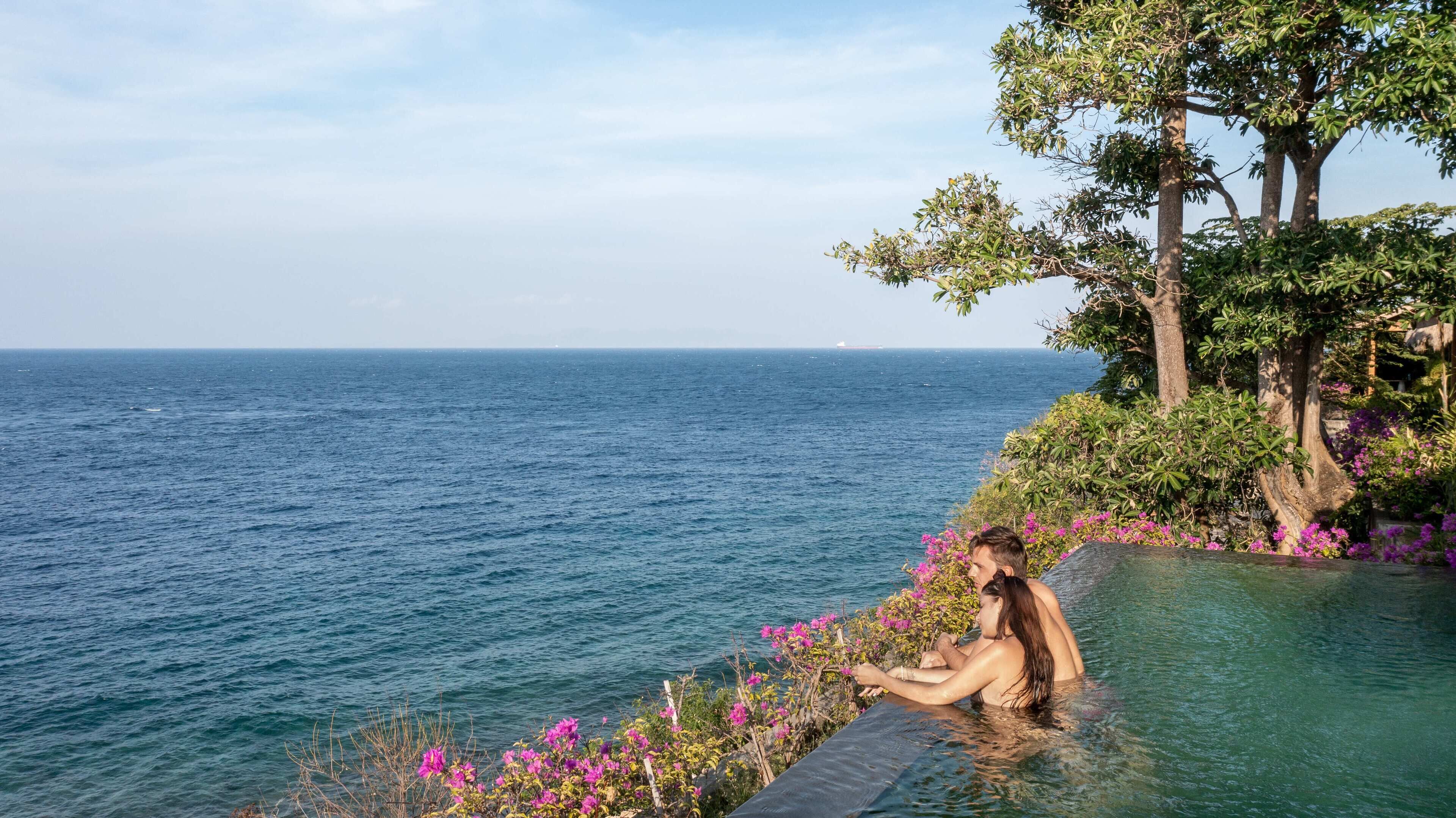 Une personne assise dans une piscine aux eaux cristallines, avec vue sur la mer et un arbre aux fleurs roses.