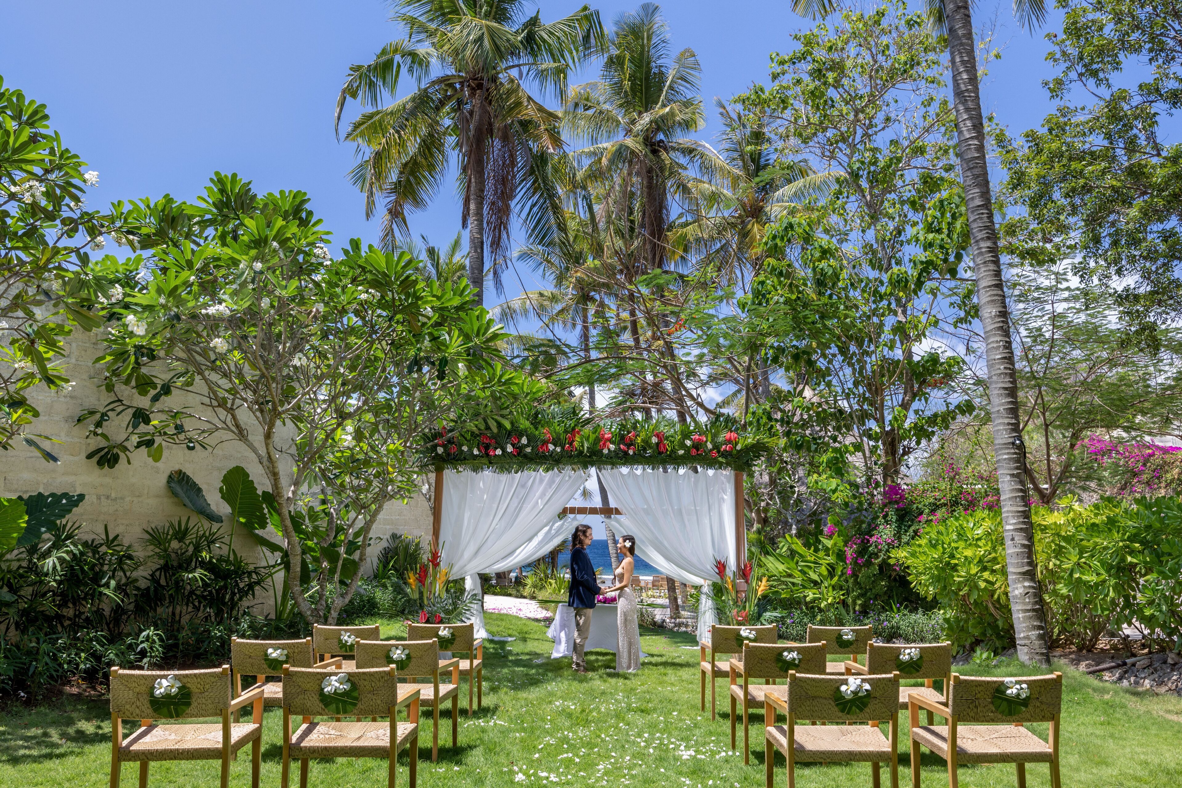 Une cérémonie de mariage en plein air, avec un dais blanc, des invités assis sur des chaises en osier et des plantes tropicales en arrière-plan.
