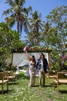 A wedding ceremony with a white canopy and floral decorations, set in a garden with palm trees and chairs.